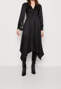 Robe en satin noir avec un décolleté en V, un corsage plissé, des manches longues et un ourlet asymétrique. Associée à des bottes noires ajustées jusqu'au genou.