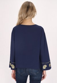 Top blu navy con vestibilità ampia, maniche a campana larghe, con ricami dorati attorno ai polsini. Tessuto morbido e scollatura rotonda semplice.