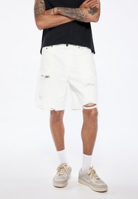 Shorts de mezclilla blanca con desgastes y dobladillos deshilachados, combinados con una camiseta negra y zapatillas neutras. Textura suave y ajuste casual.