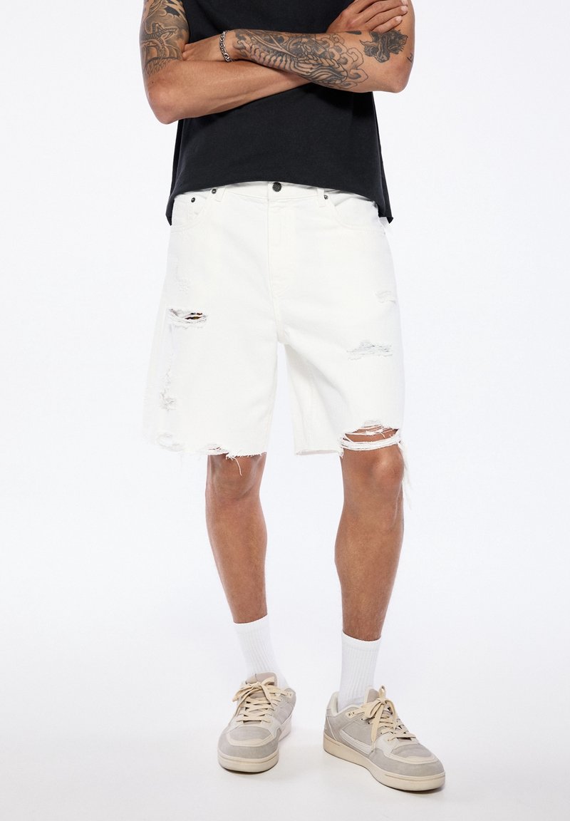 Shorts de mezclilla blanca con desgastes y dobladillos deshilachados, combinados con una camiseta negra y zapatillas neutras. Textura suave y ajuste casual.
