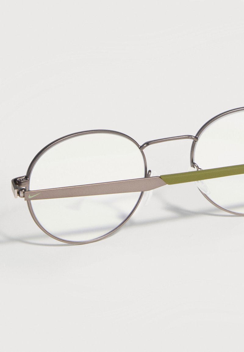 Runde Metallbrille mit dünnen silbernen Fassungen und olivgrünen Bügeln, vor einem schlichten hellen Hintergrund gezeigt.