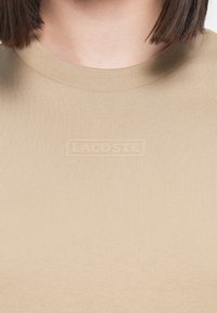 T-shirt beige a coste con scollatura rotonda e logo in rilievo discreto. Il tessuto appare morbido, offrendo una finitura testurizzata senza ulteriori motivi.