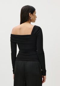 Crna dugosavna majica s asimetričnim, off-shoulder dizajnom; ima naboranu teksturu i glatku tkaninu. Nosena s crnim satenastim hlačama.
