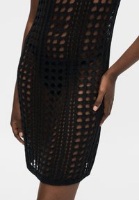 Κοντινό πλάνο ενός ατόμου που φοράει μαύρο πλεκτό φόρεμα πάνω από μαύρα εσώρουχα, με το χέρι να ακουμπάει στο ισχίο σε ουδέτερο φόντο.