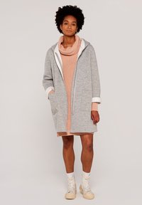 Grauer, oversized Reißverschluss-Hoodie mit weißem Innenfutter, ausgestattet mit einem großen Kragen und vorderen Taschen. Kombiniert mit einem rosa Strickpullover und cremefarbenen Stiefeln.