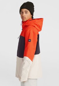 Chaqueta a bloques de color con secciones en naranja, marino y crema. Material impermeable con capucha y bolsillos con cremallera. Parche de logo negro en la manga.