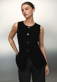 Sort fløjl, ærmeløs vest med seks metal knapper og sidelommer, der har en tætsiddende silhuet og glat tekstur. Bruges sammen med sorte bukser.