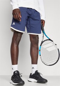Calções atléticos azul marinho com acabamento branco e logótipo da Adidas, combinados com sapatilhas pretas e meias brancas até ao tornozelo, segurando uma raquete de ténis.