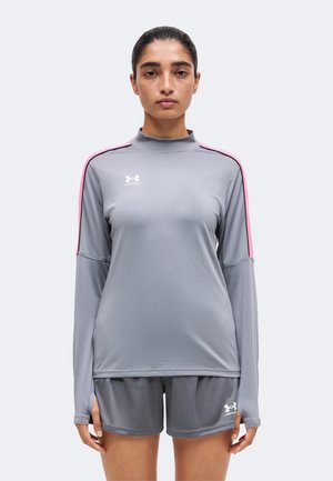 Femme portant un haut de sport gris à manches longues avec des bandes roses sur les épaules et un short gris assorti, debout face à un fond uni.
