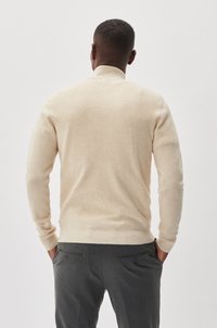 Beige kötött pulóver magas gallérral, hosszú ujjal és bordázott mandzsettával, szürke nadrággal, hátsó zsebekkel párosítva.