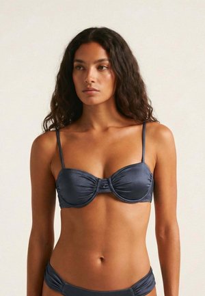 Joven mujer con cabello largo y rizado, vistiendo un bikini azul oscuro, de pie contra un fondo claro y sencillo.