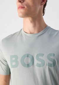 Joven con una camiseta gris clara con la palabra "BOSS" en relieve y de gran tamaño en el pecho, mirando ligeramente hacia la izquierda contra un fondo sencillo.