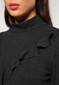 Ciemnoszary top z golfem z falbanką z przodu, wykonany z teksturowanej tkaniny, o dopasowanym kroju i subtelnym wysokim dekolcie.