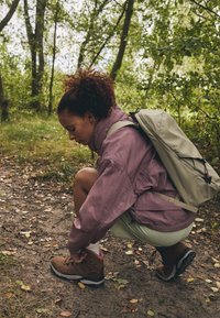Bruine wandelschoenen met roze accenten, lichtgroene shorts, paarse jas en een lichtgrijze rugzak, hurkend op een bosbodem met bladeren.