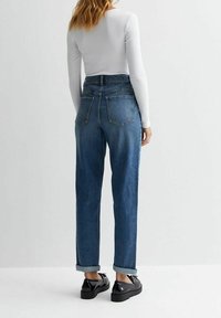 Nainen, jolla on ylhäältä hihoissa käänteet siniset korkeavyötäröiset farkut, valkoinen pitkähihaisuus paita ja mustat kiiltävät platta-kengät, seisoo yksinkertaista vaaleaa taustaa vasten.