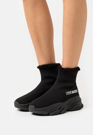 Jambes portant des baskets à chaussettes noires à la cheville avec des semelles texturées et le logo "Steve Madden" sur le côté, ornées d'un bord de manchette duveteux.