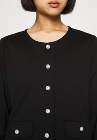 Fekete dzseki lekerekített nyakkivágással, öt fényes ezüst színű gombbal és két elülső zsebbel. Sima szövet textúra.