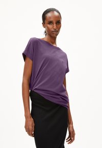 Korte paarse t-shirt met opgerolde mouwen, losse pasvorm en een klein embleem op de borst. Gecombineerd met een zwarte strakke rok.