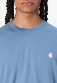 T-shirt en coton bleu clair avec un col rond et des manches courtes ; présente un petit logo blanc sur la zone du côté gauche de la poitrine.
