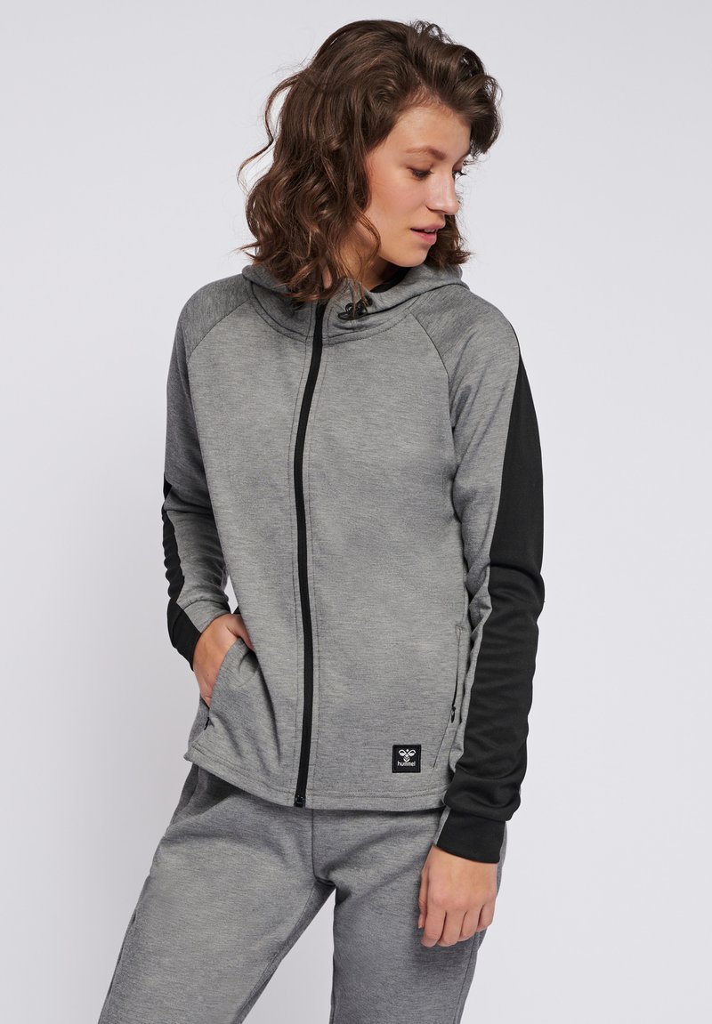 Vrouw draagt een grijze hoodie met ritssluiting en zwarte mouwen en bijpassende grijze broek, kijkt naar beneden met één hand in de zak.