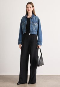 Jeansová bunda s cropped strihom, modrej farby, s náprsnými vreckami, zladená s čiernymi širokými nohavicami a čiernou koženou kabelkou. Hladká textúra.