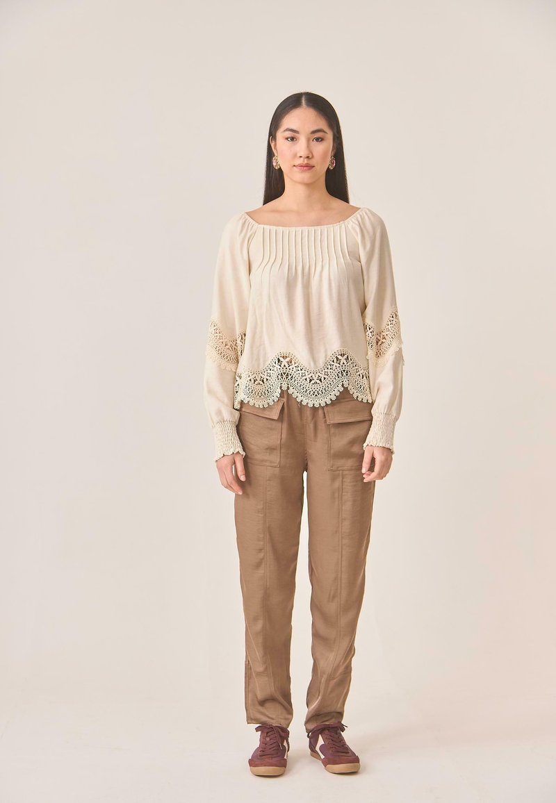 Blusa crema con tejido texturizado y borde de encaje, combinada con pantalones de pierna ancha marrones. La modelo lleva zapatillas burdeos.
