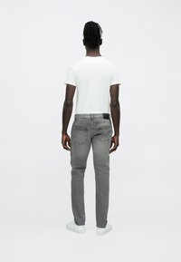 Vaqueros de mezclilla gris con un ajuste slim, bolsillos traseros y una etiqueta negra en la cinturilla, combinados con una camiseta blanca lisa y zapatillas blancas.