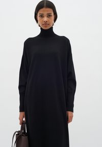 Svart polotröja klänning, oversize passform, lång ärm. Slät textur, knälängd, tillsammans med en brun handväska. Enkel, modern design.