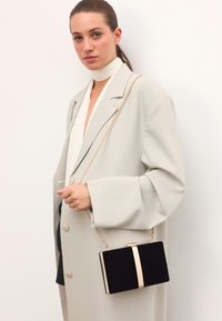 Juodas aksominis vakarėlio krepšys su auksiniu akcentu ir grandinės dirželiu, laikomas modelio, vilkinčio šviesiai pilką per didelį paltą ir kreminę palaidinę.