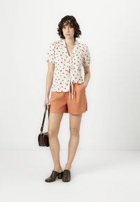 Blomstrete kortermet bluse med knytedetalj, kremfarget bakgrunn og rødt blomstermønster. Kombinert med oransje shorts og mørke perforerte klosser, holder en brun veske.