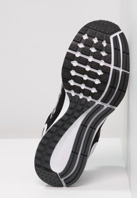 Semelle de chaussure de course noire et blanche avec un motif en caoutchouc texturé, comprenant des rainures et des crampons circulaires pour une meilleure traction et stabilité.