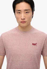 Camiseta de rayas rojas y blancas con cuello redondo y mangas cortas, que presenta un pequeño logo bordado rojo en el pecho. Textura de tela suave.