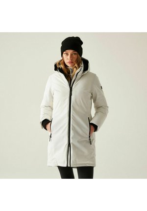 Mujer con abrigo blanco de invierno con capucha, detalles en cremalleras negras y un gorro negro, con las manos en los bolsillos, de pie frente a un fondo liso.