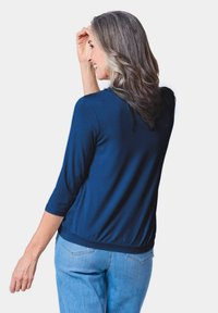 Dunkelblauer, leichter Jersey-Oberteil mit drei Viertelärmeln, gerafftem Saum, weicher Textur, getragen mit hellblauen Jeans. Rückansicht.