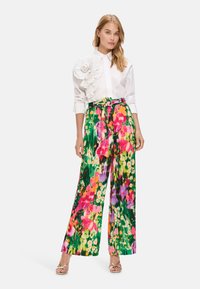 Weißes Oberteil mit strukturellem Blumenakzent, kombiniert mit lebhaften, weiten Hosen mit einem mehrfarbigen Blumenmuster. Silbersandalen.