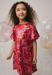 Vestito rosso con paillettes e maniche corte svolazzanti, caratterizzato da un motivo floreale in tonalità di rosso più chiare, indossato da un bambino che tiene un palloncino.