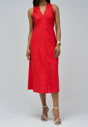 Kobieta w czerwoną, bezrękawnikową sukienkę midi z dekoltem w kształcie litery V oraz białymi sandałami na wysokim obcasie ze smyczkami, stojąca na jednolitym tle.