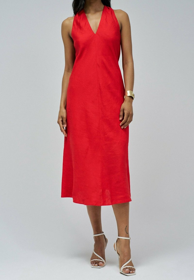 Femme portant une robe midi rouge sans manches avec un col en V et des sandales blanches à talons hauts à lanières, debout devant un fond uni.