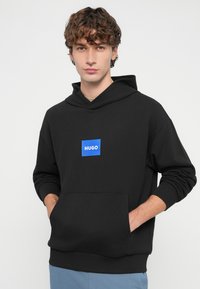 Mladý muž s kučeravými vlasmi, oblečený v čiernej kapucni s modrým logom "HUGO", ruky v predných vreckách, a svetlomodré nohavice.