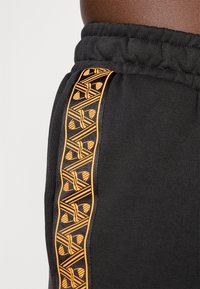 Czarne dresy z elastycznym pasem mają pomarańczowy, geometryczny wzór wzdłuż boku. Miękka, gładka struktura materiału.