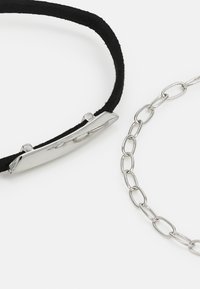 Bracelet en suède noir avec une plaque en métal argenté et bracelet en chaîne argentée avec des maillons ovales, tous deux présentant des textures lisses et des lignes épurées.