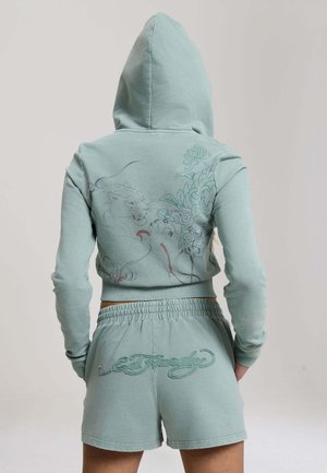 Person iført en lyseblå hættetrøje og matchende shorts med en detaljeret grafik af en brølende tiger og blomsterdesign på ryggen.