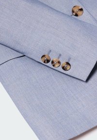 Manica di blazer su misura azzurro chiaro con tre bottoni marroni lucidi, tessuto testurizzato che mostra un sottile motivo di trama. Tessuto di rivestimento e cuciture visibili.