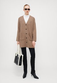 Brun, strikket cardigan med V-hals, frontknapper og to lommer; kombinert med svarte bukser og en svart-hvitt totebag.