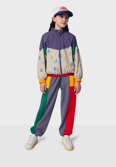 Młoda osoba stojąca, ubrana w fioletową, beżową i wielokolorową kurtkę z literami, kolorowe spodnie w zielonym, fioletowym, czerwonym i żółtym kolorze, białe sneakersy oraz czapkę.