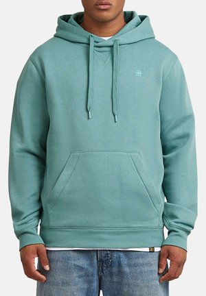 Homme portant un sweat à capuche à enfiler de couleur bleu-vert clair avec une poche avant et des cordons, associé à un jean bleu et un sous-vêtement blanc visible à l'ourlet.