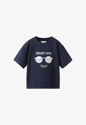 T-shirt bleu marine avec le texte "JOURS DE VACANCES : NAGE, RIRE, EXPLORER !" et un graphisme de lunettes de soleil montrant une scène de plage et de coucher de soleil.