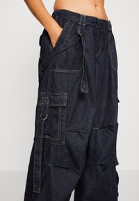 Dark denim cargo pants with multiple pockets, drawstring waist, and contrasting stitching. Texture appears sturdy; design is oversized and relaxed.