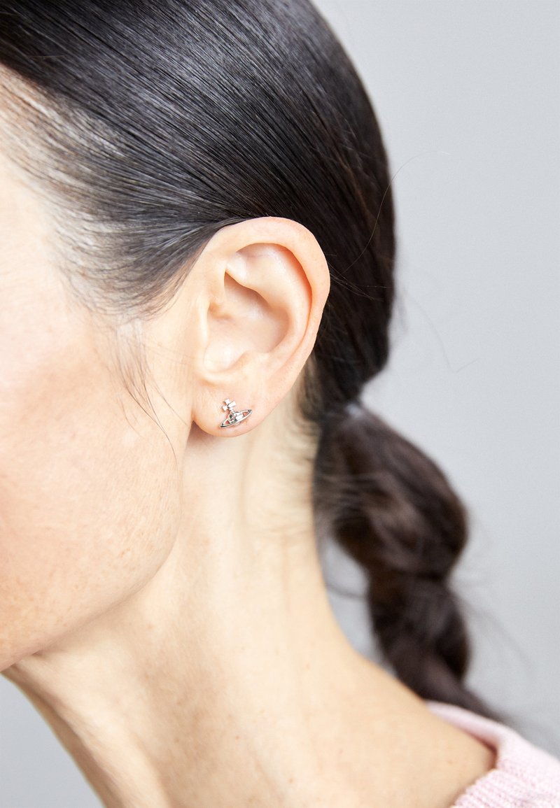 Woman with dark braided hair wearing small silver orb-shaped stud earring on her left ear against a plain light background.