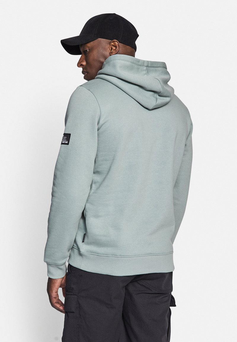 Hellgrauer Kapuzenpullover mit Kängurutasche, gerippten Bündchen und Saum. Mit einem Logopatch am linken Ärmel. Kombiniert mit dunklen Cargo-Hosen.
